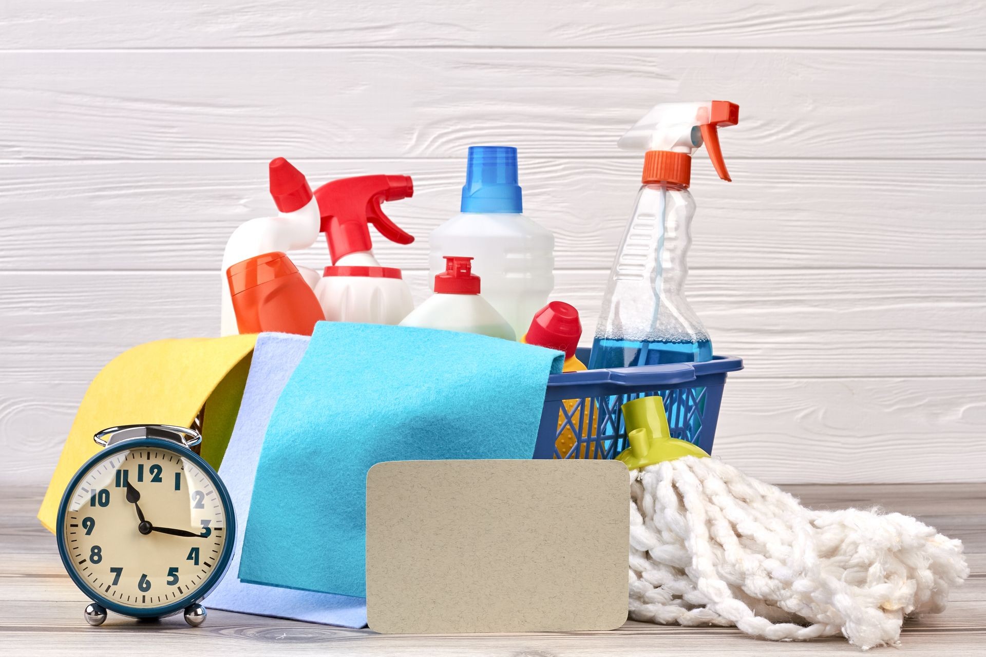 Variety of cleaning supplies on wooden background. Plastic basket with cleaning products, alarm clock and paper card. Domestic cleaning company.